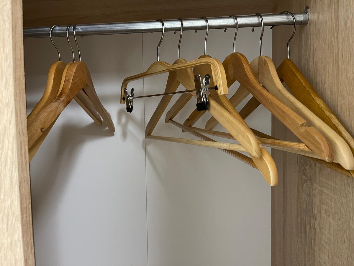 A wooden wardrobe features a hanging rod with several wooden hangers displayed. The interior is equipped with a neutral backdrop, providing ample space for clothing storage. The arrangement suggests organization and convenience for guests during their stay.