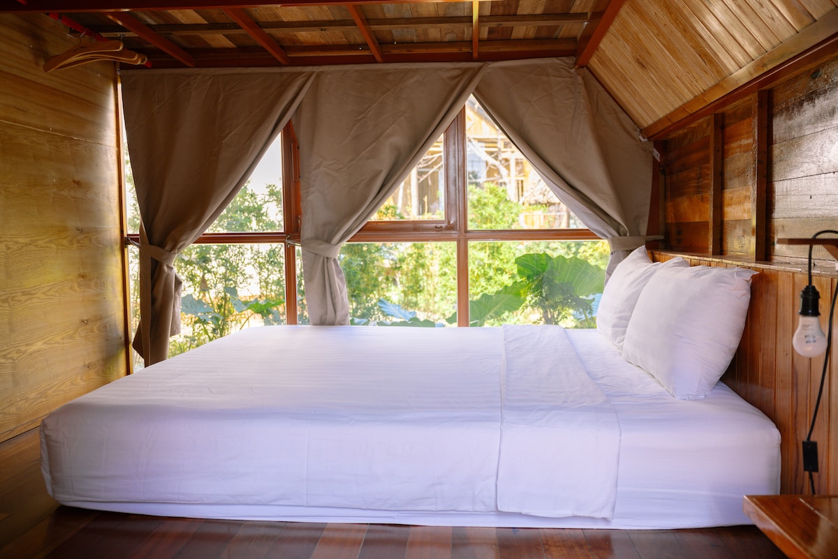 A serene sleeping area is highlighted by large windows with soft curtains, allowing natural light to fill the space. A spacious bed is dressed with crisp white linens, offering a peaceful environment amid wooden walls and charming natural surroundings.