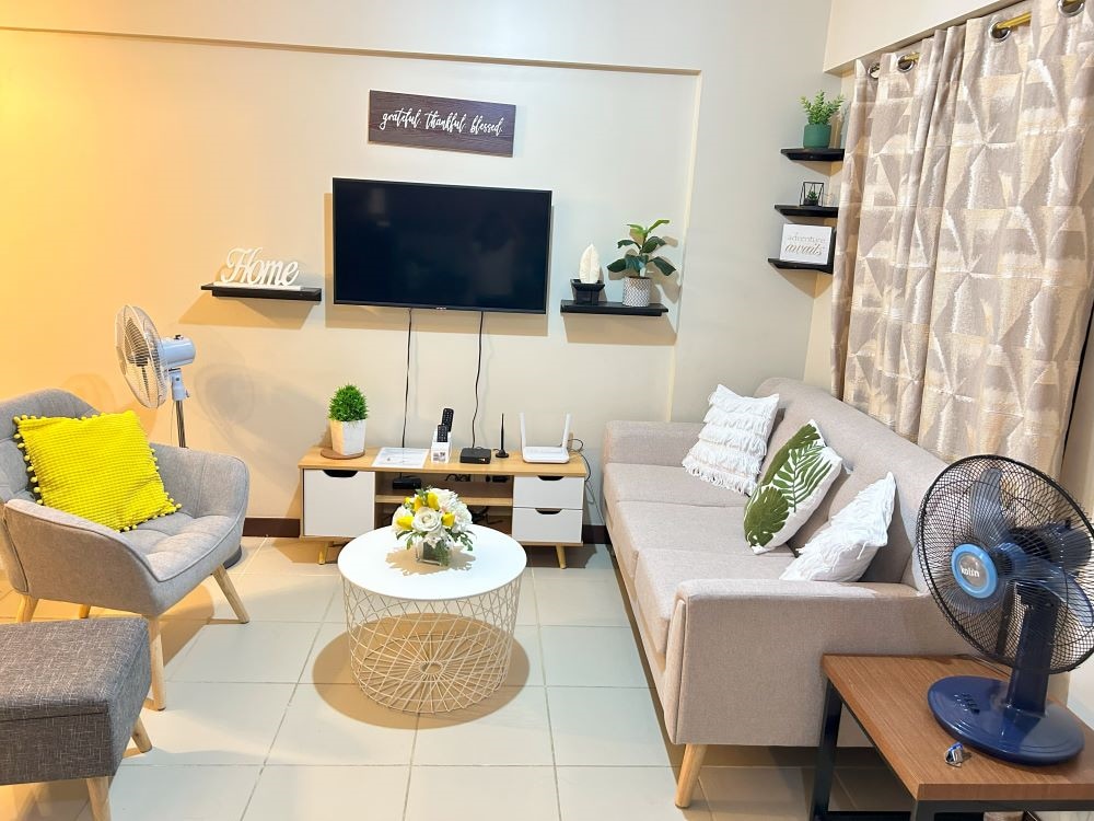 A seating area is presented with a modern light gray sofa and a cozy accent chair. A round white coffee table is topped with a small floral arrangement. A wall-mounted TV is positioned above a low cabinet, flanked by decorative items and a fan.