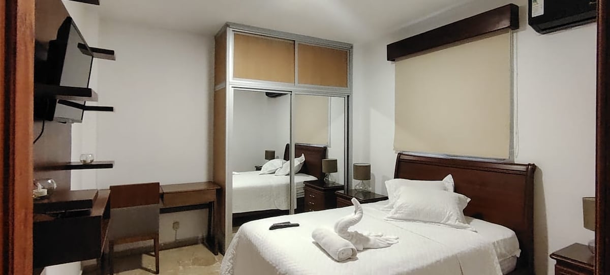 A well-appointed room features a spacious bed with white bedding and decorative towels arranged in a swan shape. A mirrored wardrobe reflects the surroundings, while a desk and lamp provide workspace. Natural light enters through a covered window, enhancing the room's comfort.