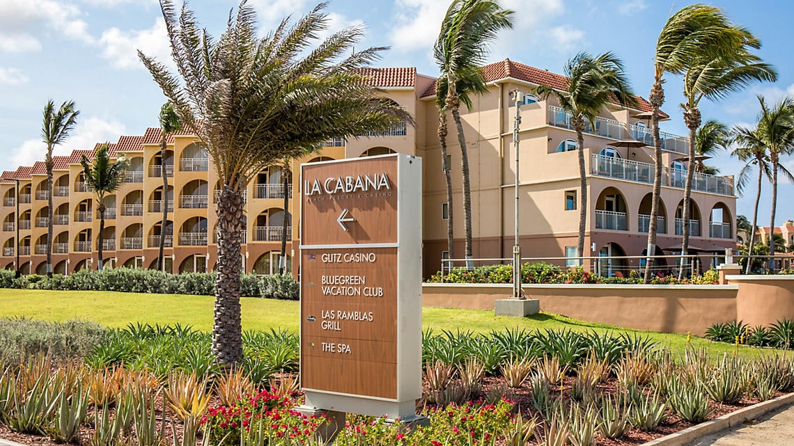 The resort's exterior features a multi-story building with balconies overlooking landscaped gardens. A sign indicates the direction to various amenities, including the casino and spa. Palm trees sway gently in the breeze, complementing the tropical atmosphere of the surroundings.