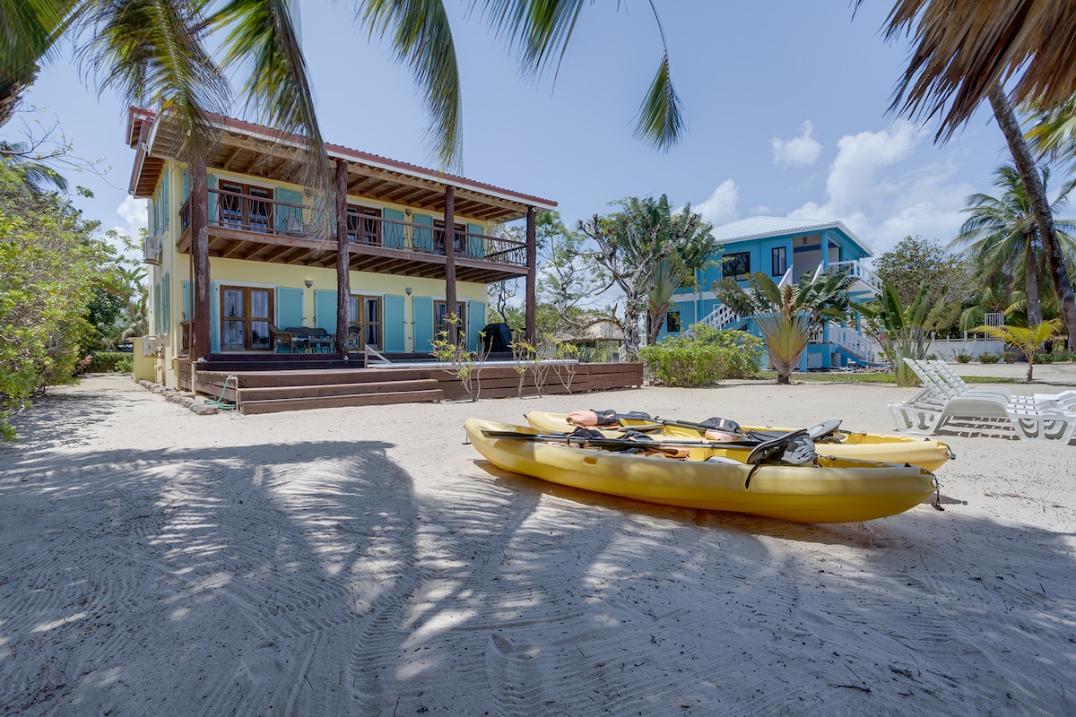 A view of the beachfront property features two levels with a spacious outdoor area. Two kayaks are positioned in the soft sand, while palm trees provide shade. The house's vibrant colors enhance its charm against a backdrop of clear blue skies.