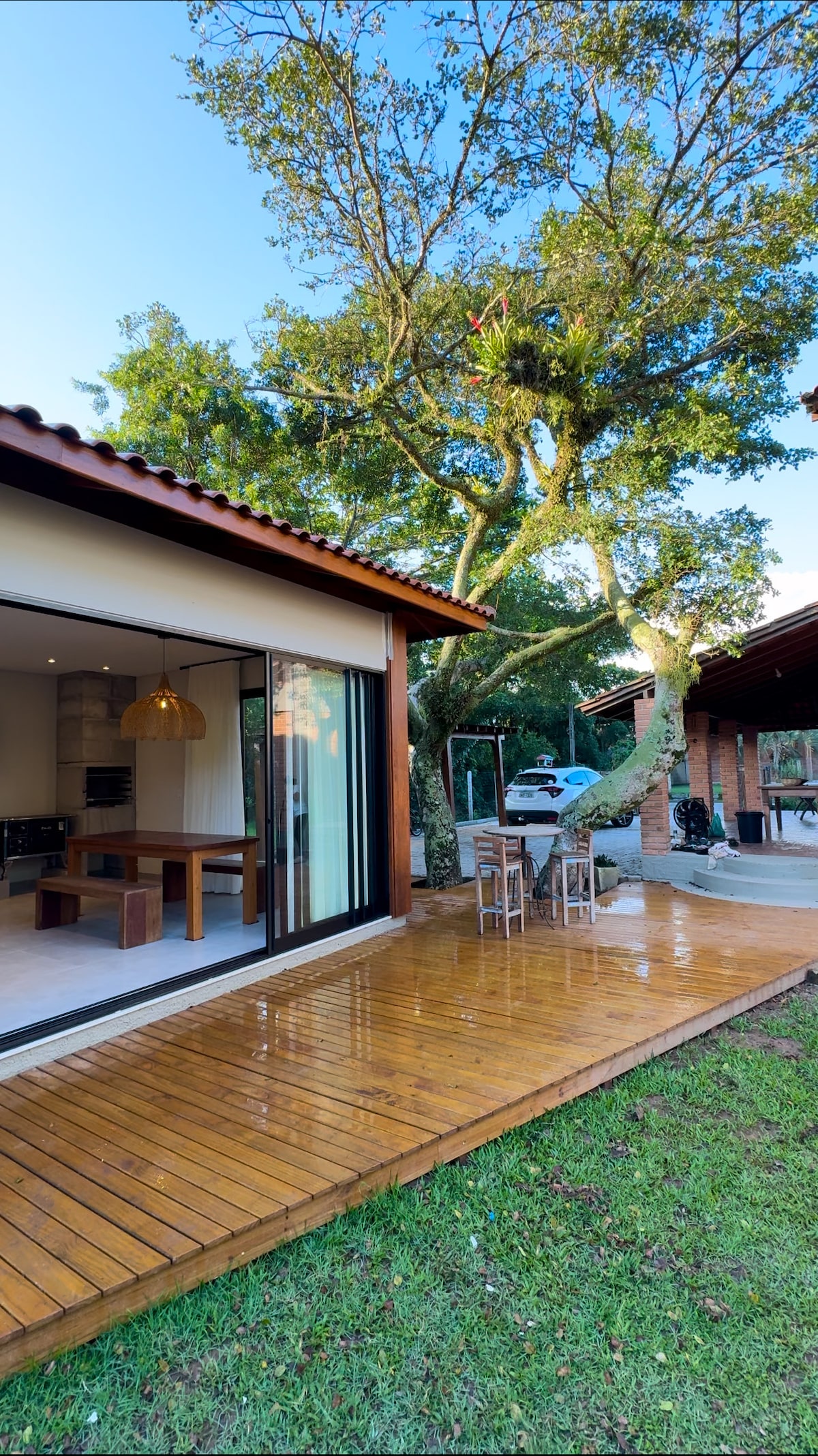 A spacious wooden deck extends from the house, surrounded by lush green grass. Large glass doors lead into a modern kitchen area, featuring a stylish light fixture above the dining table. A tree provides a natural shade, adding to the serene outdoor environment.