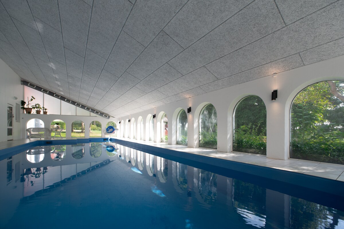 An indoor swimming pool is featured, characterized by an expansive, smooth surface reflecting light. Arched windows line the walls, providing views of the lush greenery outside. Subtle lighting adds to the serene ambiance of this tranquil space.