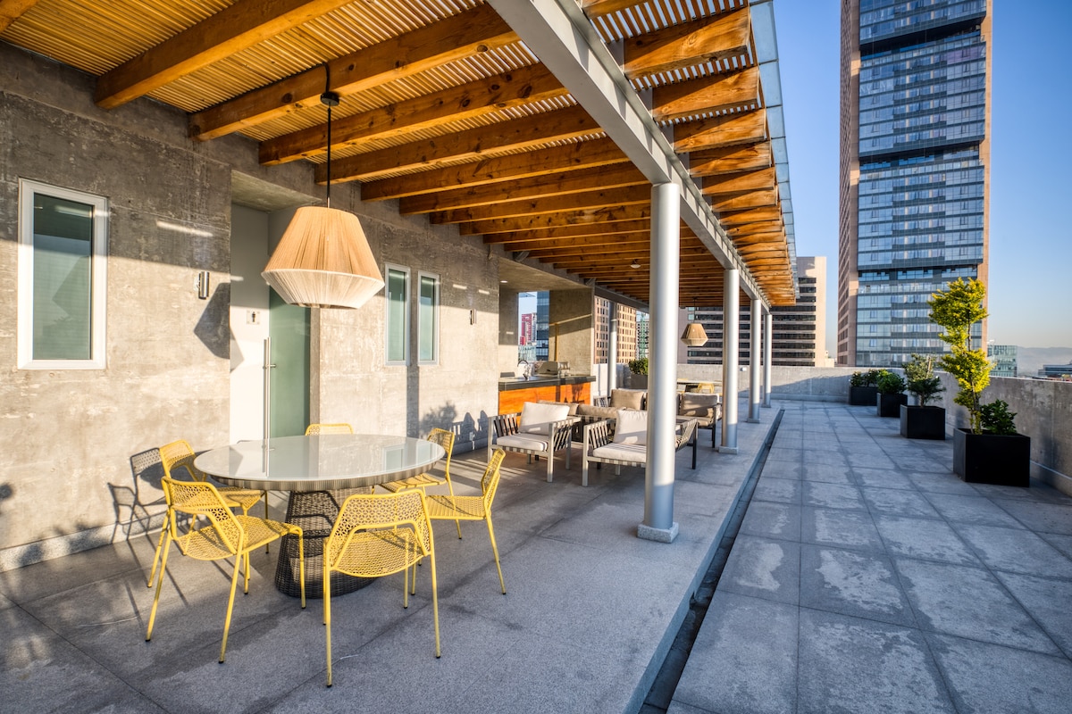 A spacious rooftop area features a round dining table surrounded by yellow chairs, ideal for outdoor meals. The wooden ceiling adds an organic touch, while modern buildings create a dynamic backdrop. Potted plants are placed along the edges, contributing to the inviting atmosphere.