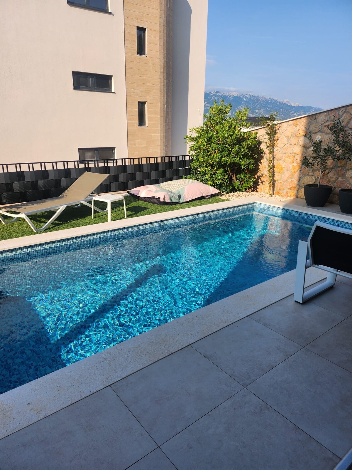 A private pool is surrounded by mosaic tiles, reflecting the clear blue water. Lounge chairs are positioned beside the pool, with a soft area of turf providing a contrast. Nearby, potted plants add greenery, while a light-colored wall offers a backdrop of warmth.