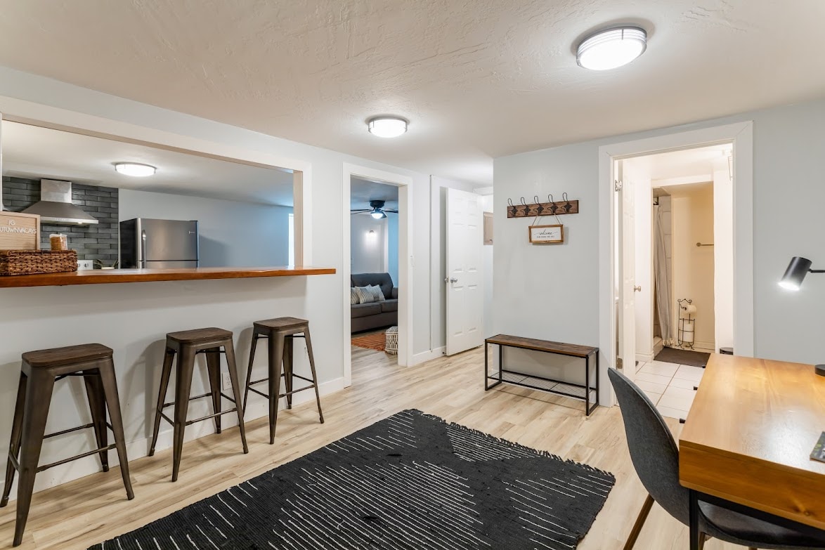An open living area showcases a modern design with light wood flooring. A rustic wooden bar separates the kitchen from the living space, complemented by three high stools. A dark rug anchors the space, while a desk and chair are positioned near a door leading to additional areas.