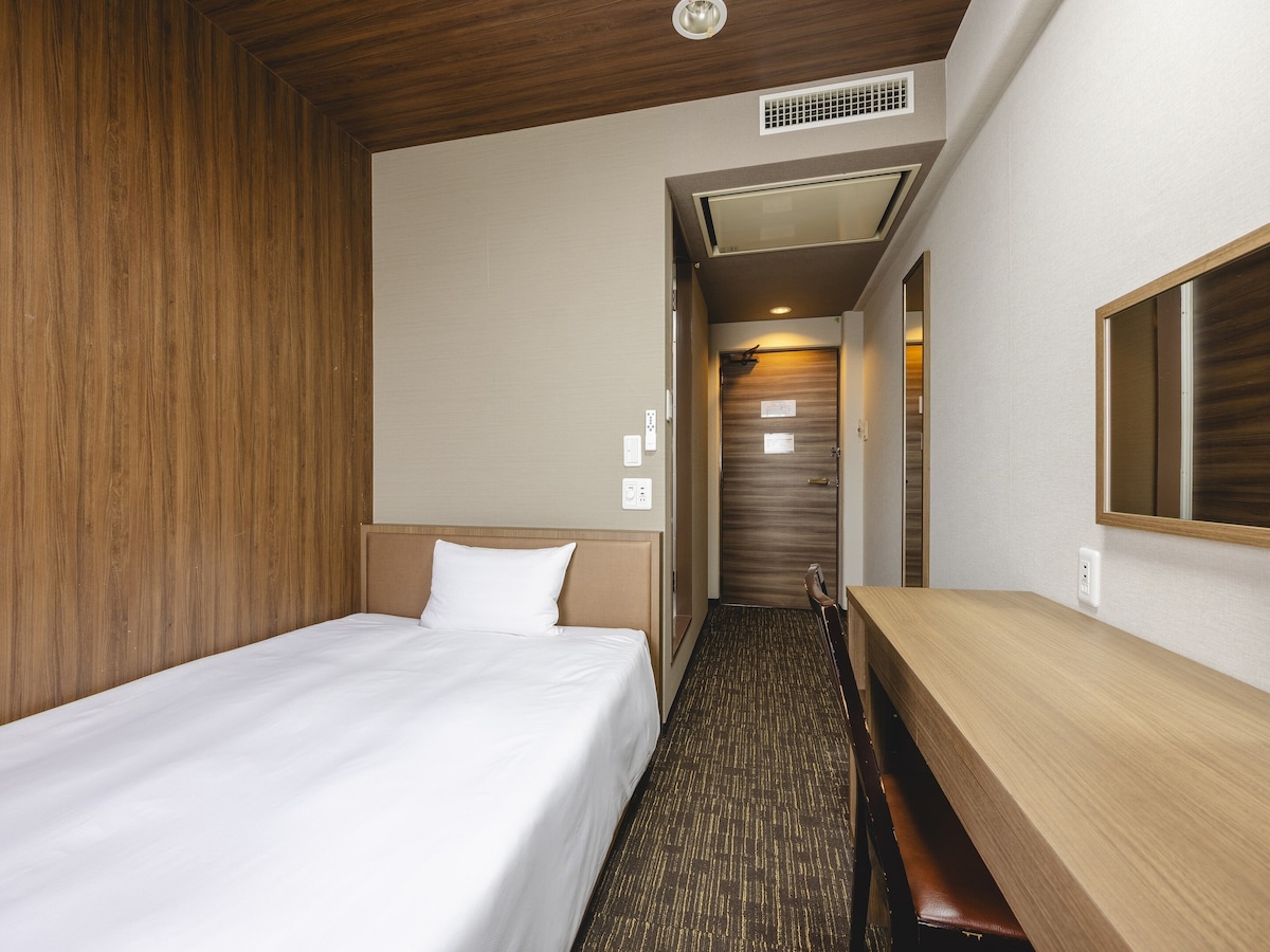 A cozy single room is furnished with a spacious bed and a wooden desk. The walls are adorned with natural wood paneling, creating a warm ambiance. A well-lit area includes a mirror above the desk, and an entrance door leads to the hallway.
