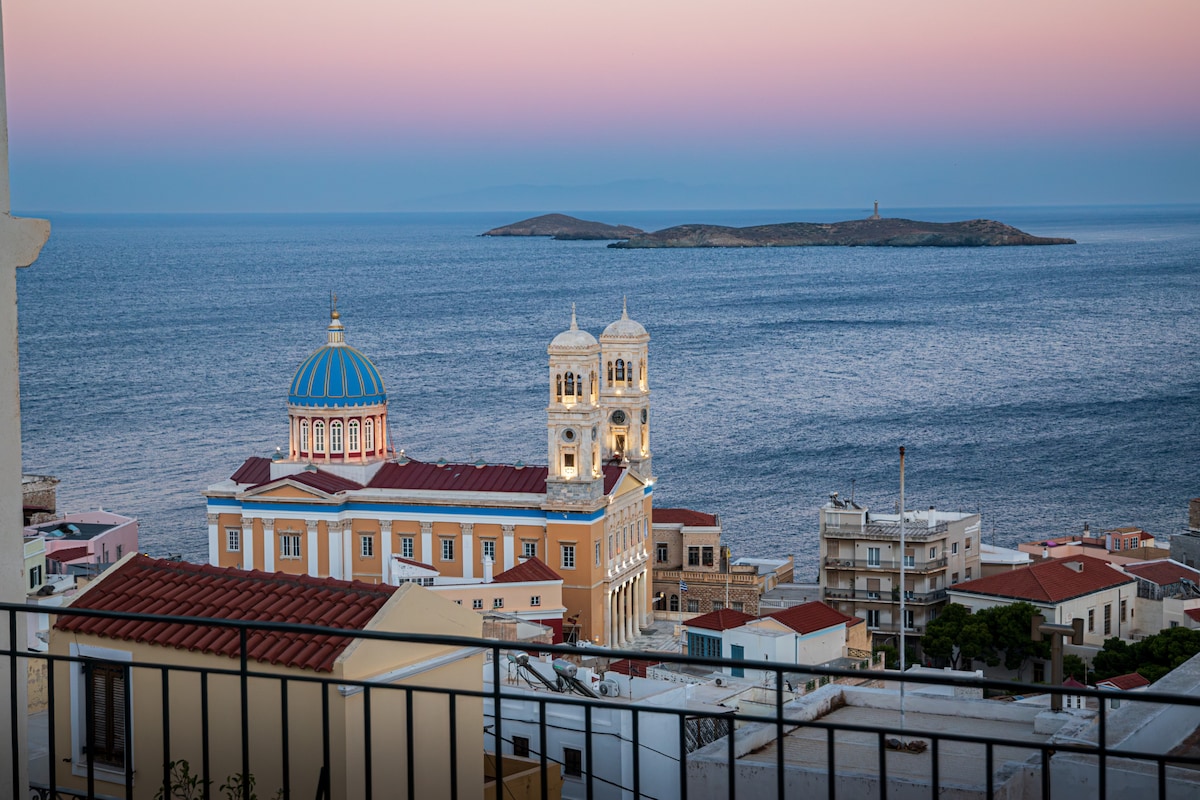 A panoramic view showcases the Aegean Sea, with gentle waves reflecting soft hues of twilight. The image features a prominent building with a blue dome, surrounded by colorful structures and distant islands, creating a serene backdrop of Ermoupoli.