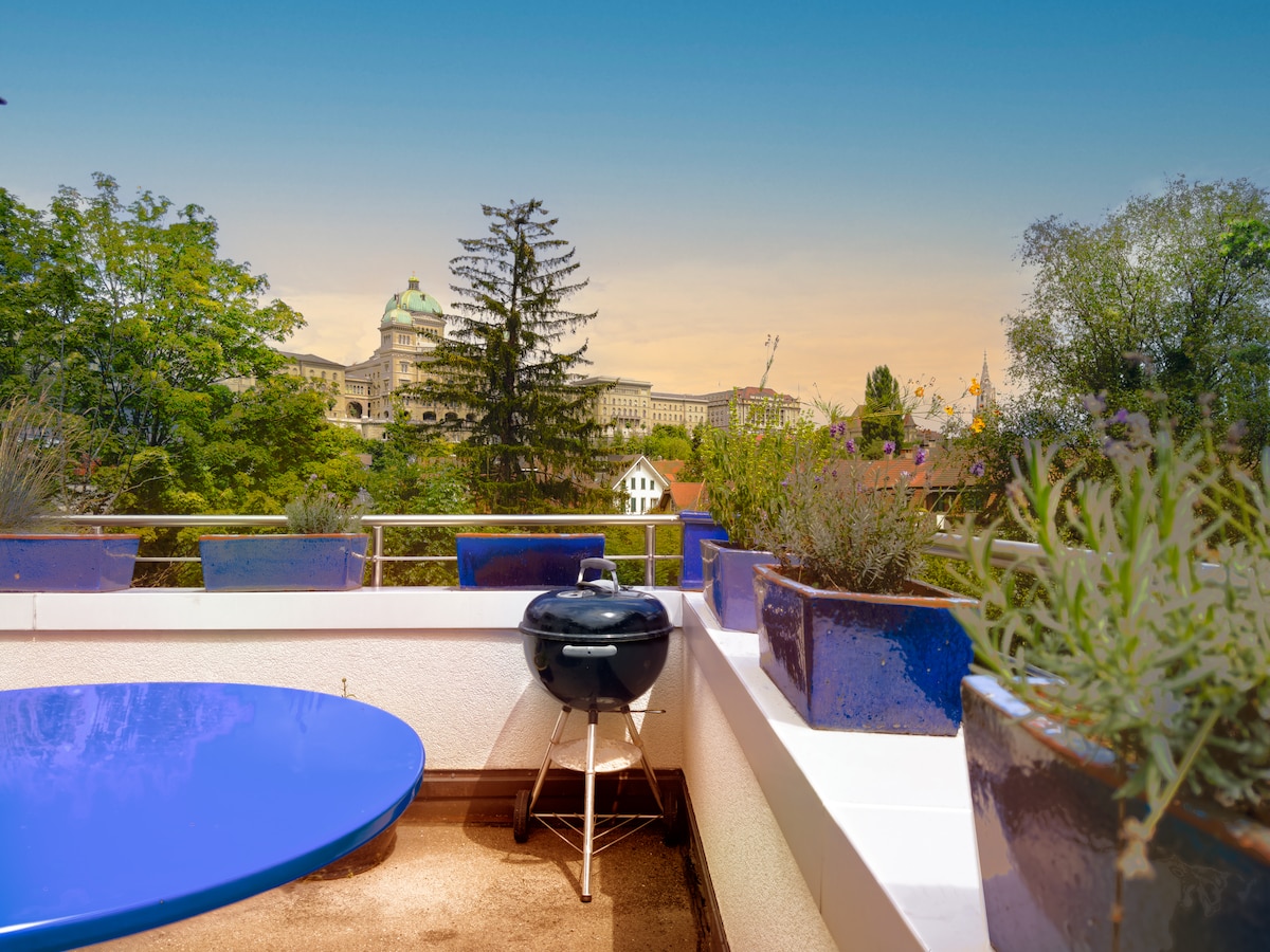 A terrace is visible, featuring a round blue table and a small charcoal grill. Surrounding plants are lush and vibrant, providing a natural setting. In the background, a view of the historic buildings of the old town of Bern can be seen against a clear sky.