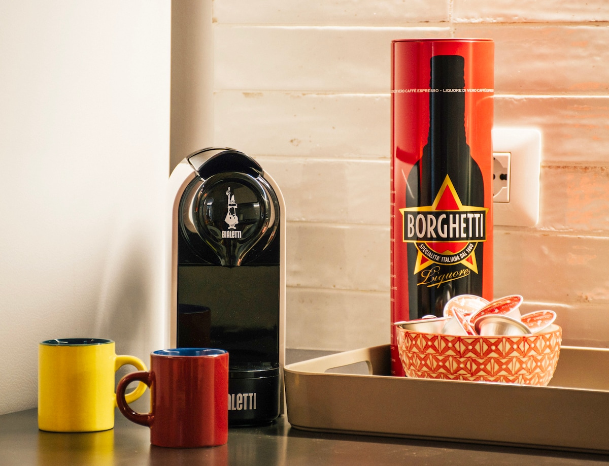 A modern coffee station features a sleek coffee machine beside colorful ceramic mugs in yellow and blue. A decorative bowl holds coffee capsules, while a red canister of liqueur adds a pop of color to the arrangement. The neutral backdrop highlights the vibrant items.