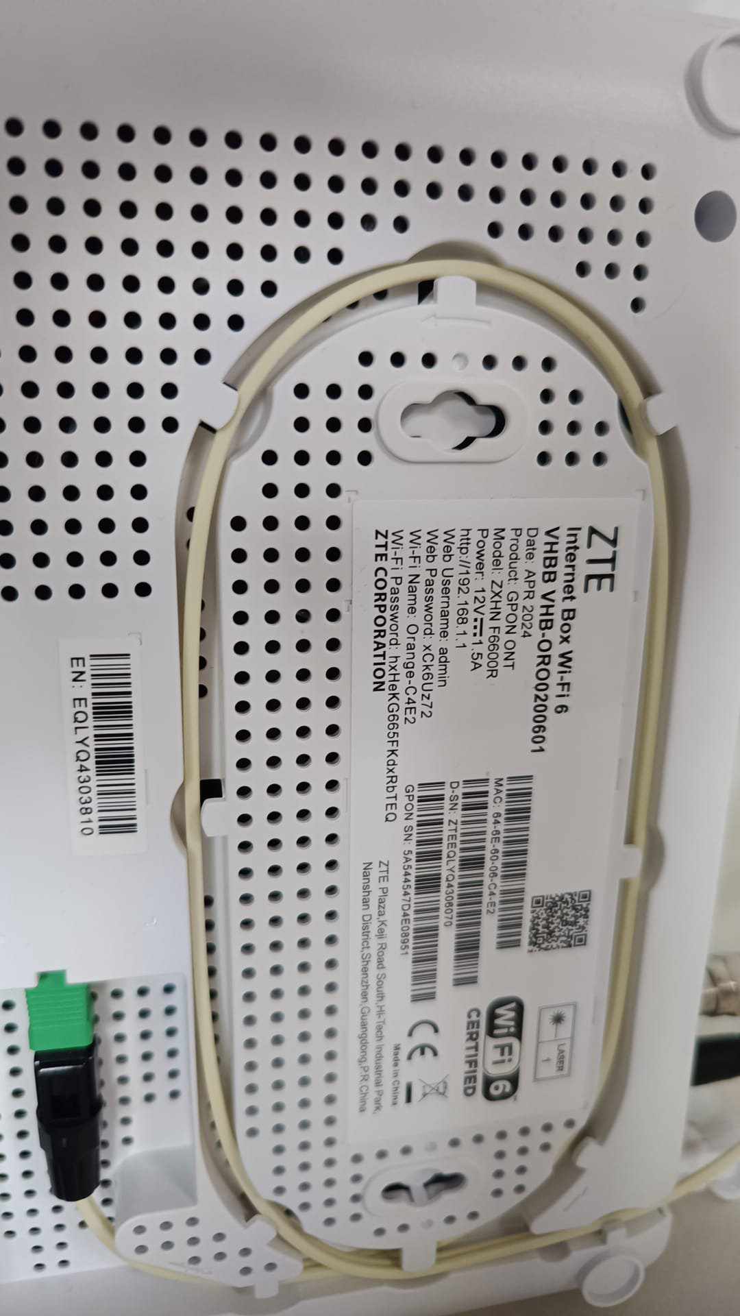 A modern internet router is mounted on a wall, featuring a white casing with ventilation holes. The back displays a series of labels and ports, indicating connectivity options. A green cable is attached, suggesting it is linked to a network source.