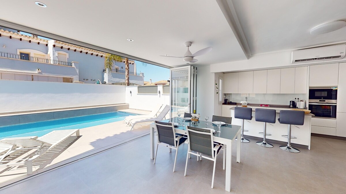 A bright dining area features a sleek glass table surrounded by modern chairs. Large sliding doors invite natural light and offer a view of the private pool and sun loungers outside. An open layout connects the space to the contemporary kitchen.