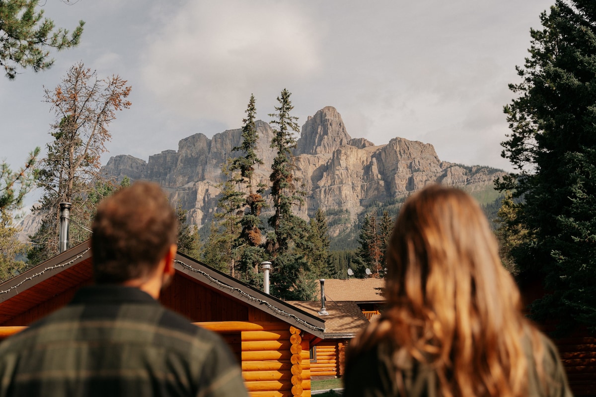 Two guests are standing outside, facing a dramatic mountain range surrounded by tall pine trees. The rustic chalets are visible in the foreground, showcasing their wooden construction and natural setting against the backdrop of rugged cliffs and a cloudy sky.
