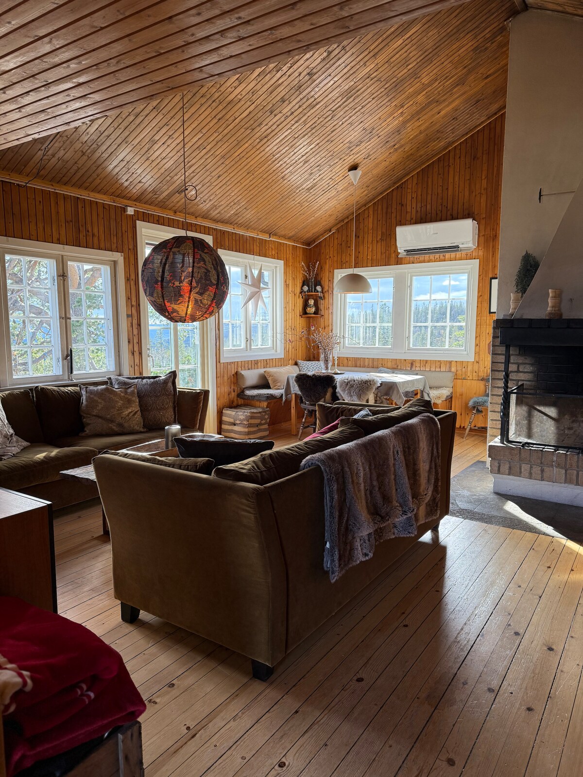 A welcoming living area features high wooden ceilings with a natural finish. Comfortable seating, including plush sofas, surrounds a fireplace. Bright light streams through multiple windows, enhancing the warm atmosphere. A decorative pendant light hangs centrally, complementing the rustic wooden decor throughout the space.