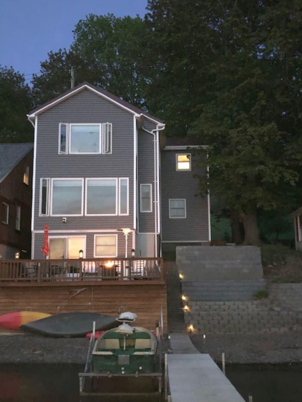 The exterior view of a three-story house is illuminated by soft lighting, showcasing large windows on the main floor. The adjoining deck features outdoor seating and is surrounded by trees, while a dock is visible in the foreground, indicating proximity to the water.