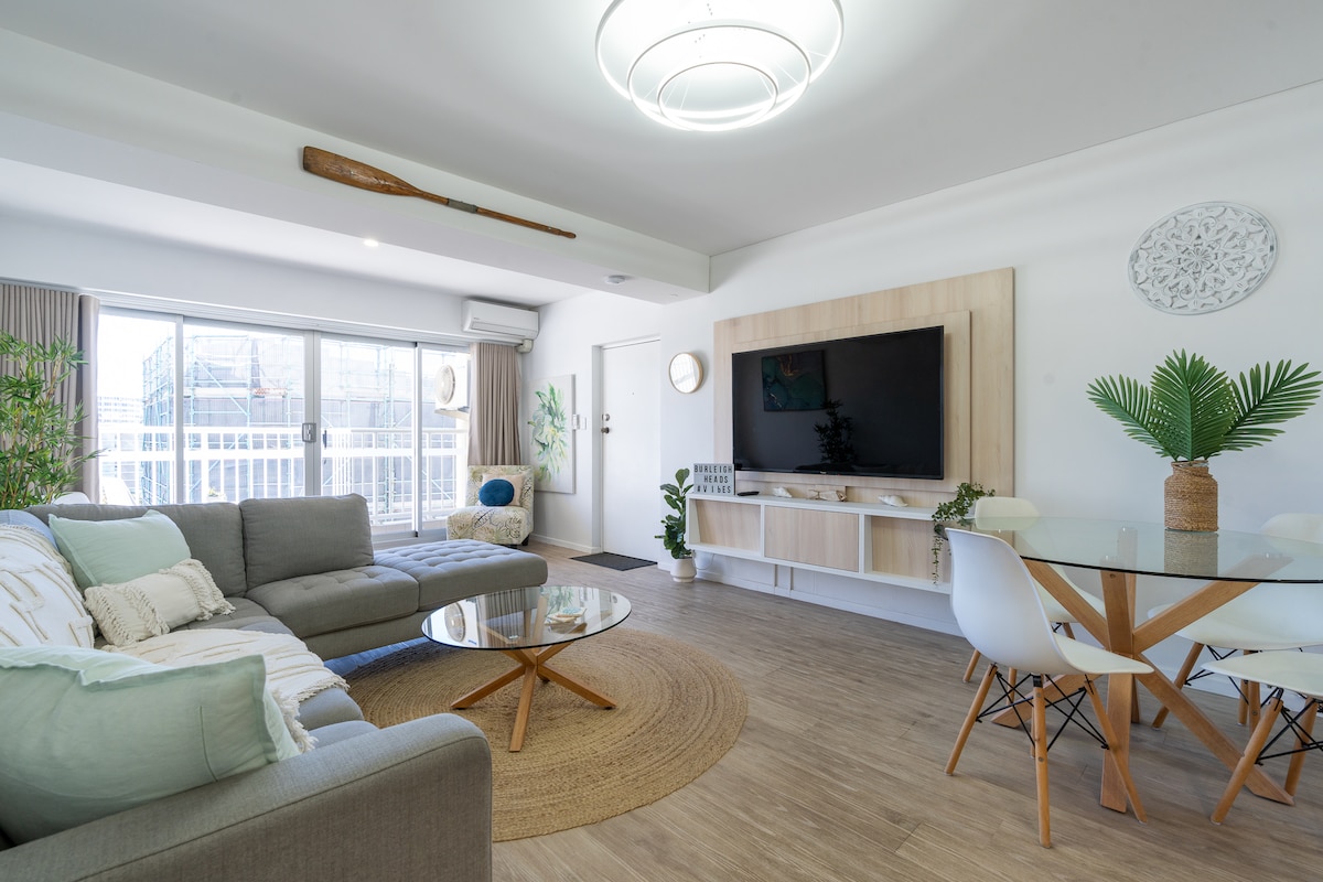 A modern living area features a comfortable gray couch with decorative pillows and a round coffee table atop a woven rug. A wall-mounted television is central to a light wooden entertainment unit. A dining table with white chairs is positioned next to large glass doors leading to a balcony.