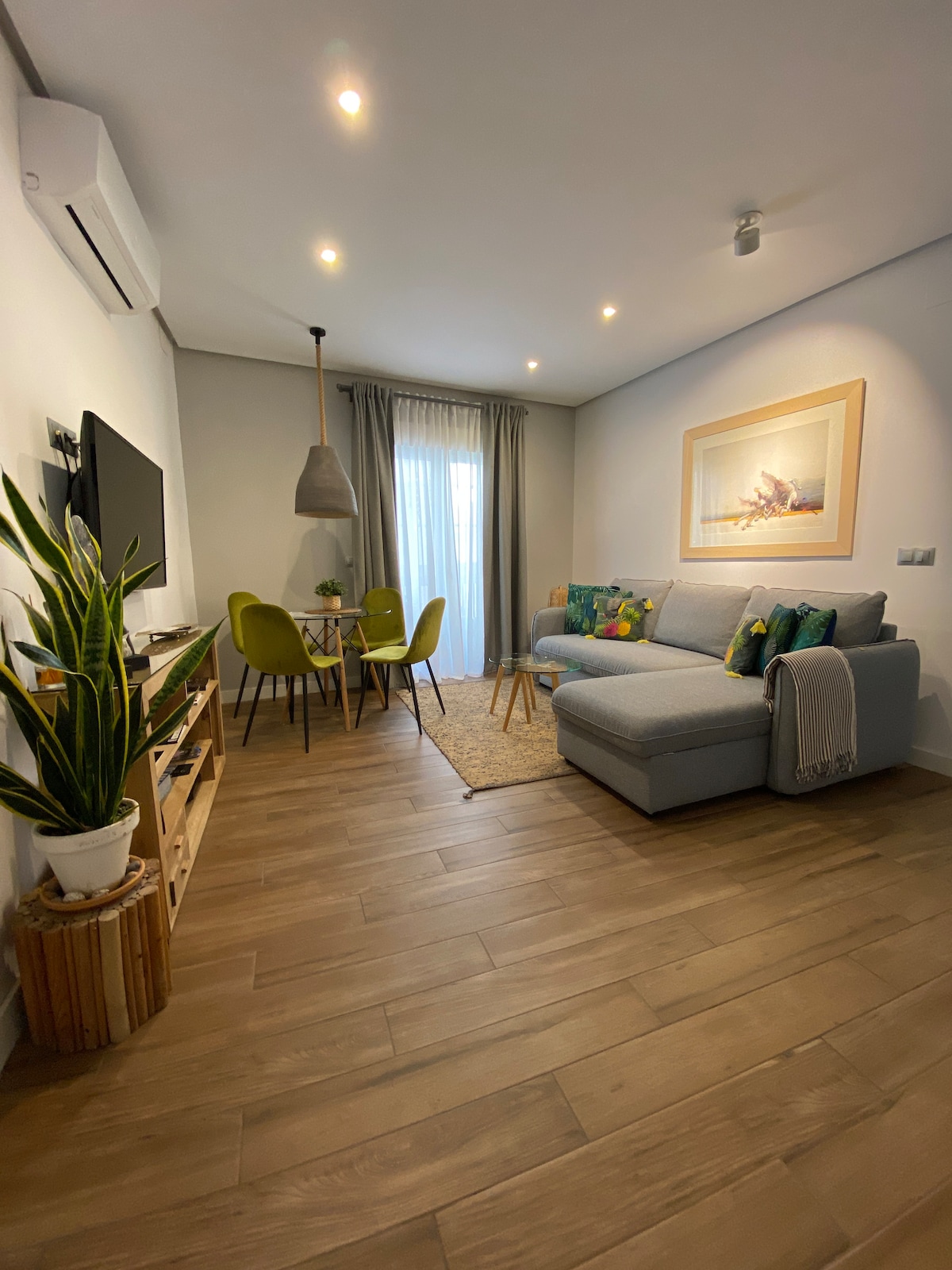 A comfortable living space is visible, featuring a light blue sectional sofa with decorative pillows. A dining area includes a round table with four green chairs. Warm wood flooring is complemented by a natural fiber rug. Large windows allow ample natural light, enhancing the overall ambiance.