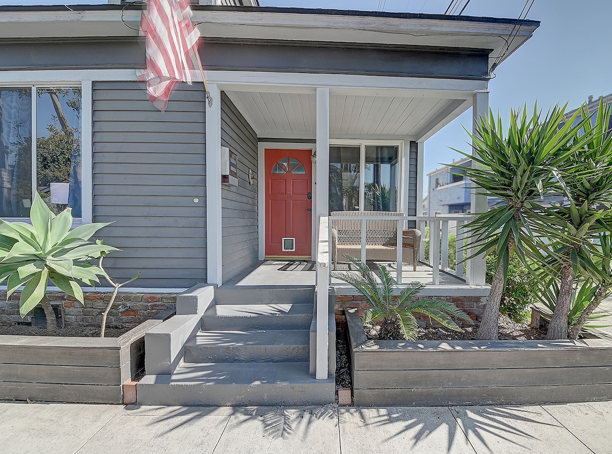 The front porch features a bright red door and a comfortable seating area. Potted plants and tropical landscaping enhance the entrance, while the walkway is flanked by low wooden planters. An American flag gently waves overhead, adding a touch of local charm.