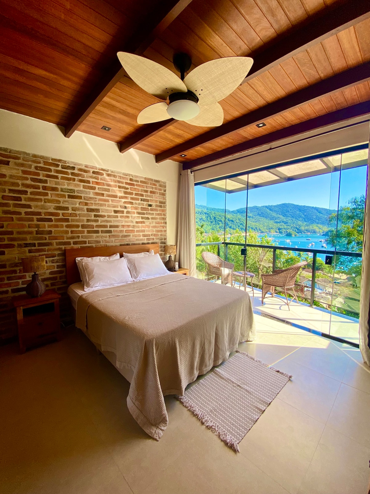 A bedroom features a spacious bed with white linens, complemented by a textured blanket. Large glass doors open to a balcony with seating, offering expansive views of the surrounding nature and mountains. The room is adorned with wooden accents and a ceiling fan.