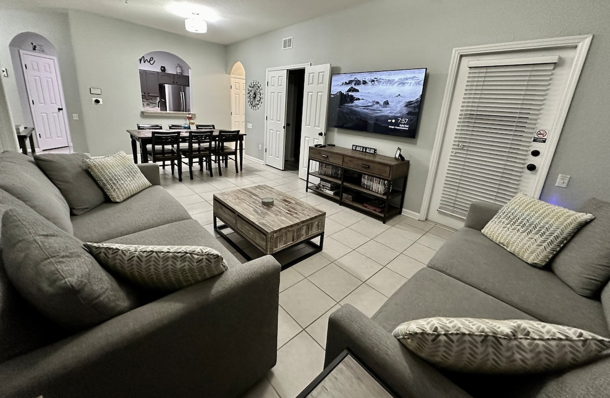 The living area features spacious gray sofas arranged around a wooden coffee table. A large wall-mounted TV displays scenic imagery. A dining area with a dark table and chairs can be seen in the background, along with a doorway leading to the kitchen.