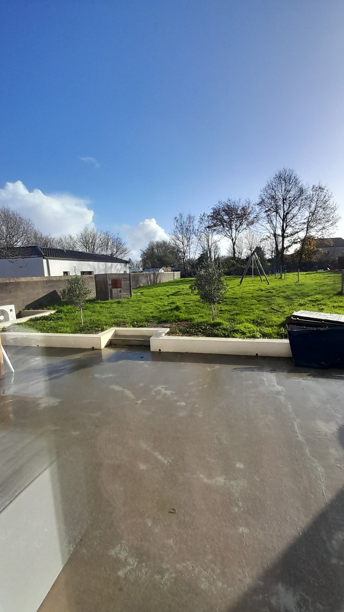 A spacious garden area is depicted, featuring a well-maintained lawn and several young trees. A play structure is visible in the background, and the sky is clear with a few scattered clouds. A portion of the terrace surface is seen in the foreground.