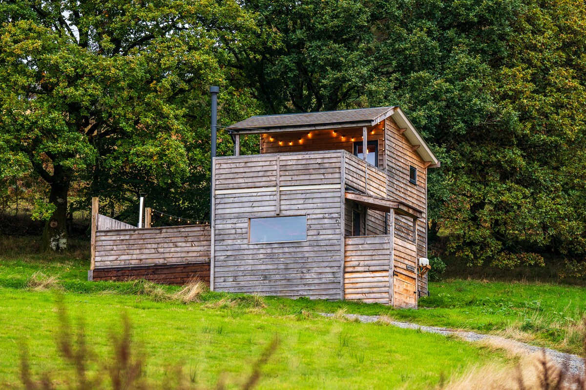 A wooden log cabin is set within a lush green landscape, elevated slightly to offer scenic views. The structure features a large deck with string lights, and a prominent window is visible on the front, reflecting the surrounding natural beauty.