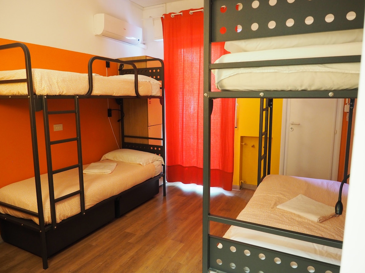 A bright room is fitted with two sets of bunk beds, providing ample sleeping space. An orange accent wall complements the warm tones of the bedding. Natural light enters through a red curtain, while a door in the background leads to the rest of the property.
