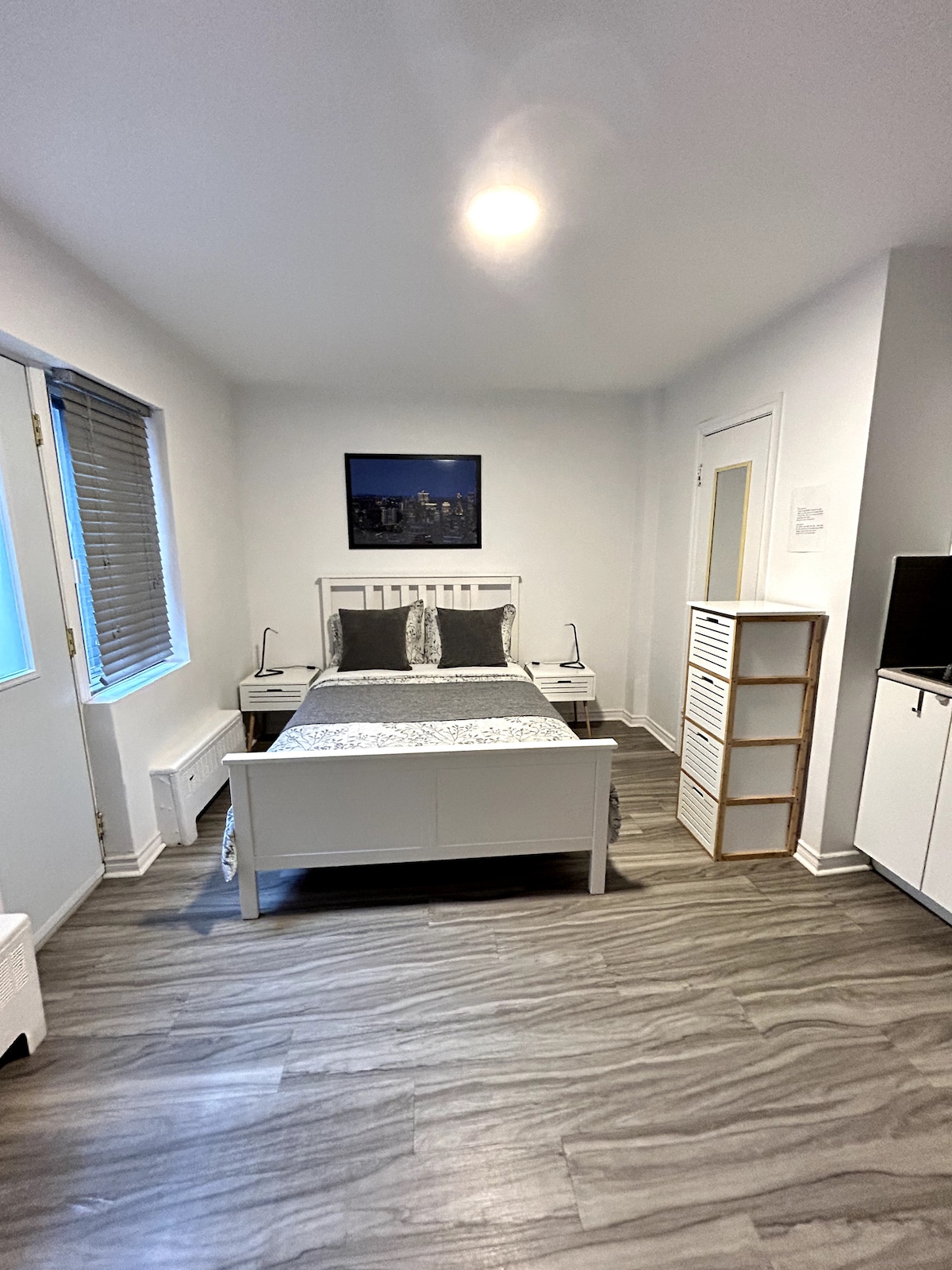 A bright studio features a spacious bed neatly made with gray linens and decorative pillows. Natural light enters through two windows, which are partially covered with blinds. A simple wooden dresser and a TV stand are positioned against the walls, complementing the warm wood flooring.