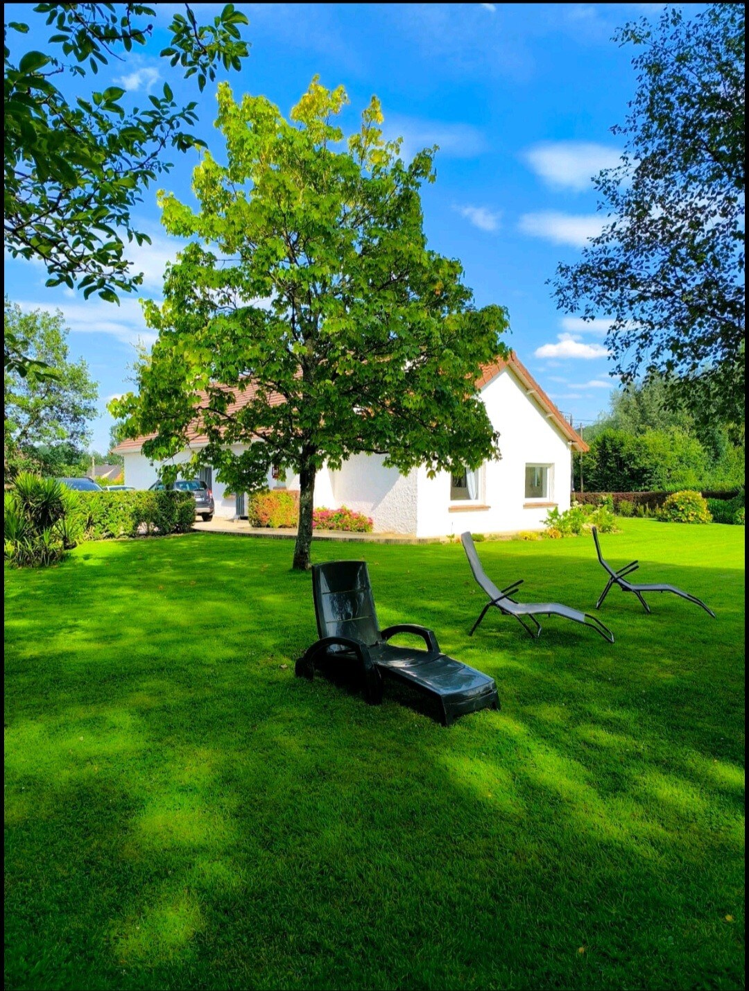 A well-maintained garden features a lush green lawn, complemented by a lounging chair and a reclining chair placed on the grass. In the background, a charming white house is surrounded by trees and colorful flower beds, all under a clear blue sky.