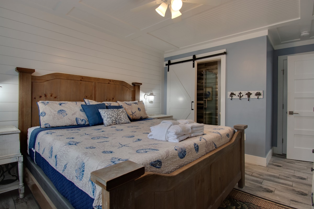 A spacious bedroom features a wooden king-sized bed with a light blue and white seashell-patterned quilt. Two bedside lamps provide illumination, and a sliding barn door leading to a bathroom adds rustic charm. Light wood accents and a cool-toned wall create a calming atmosphere.