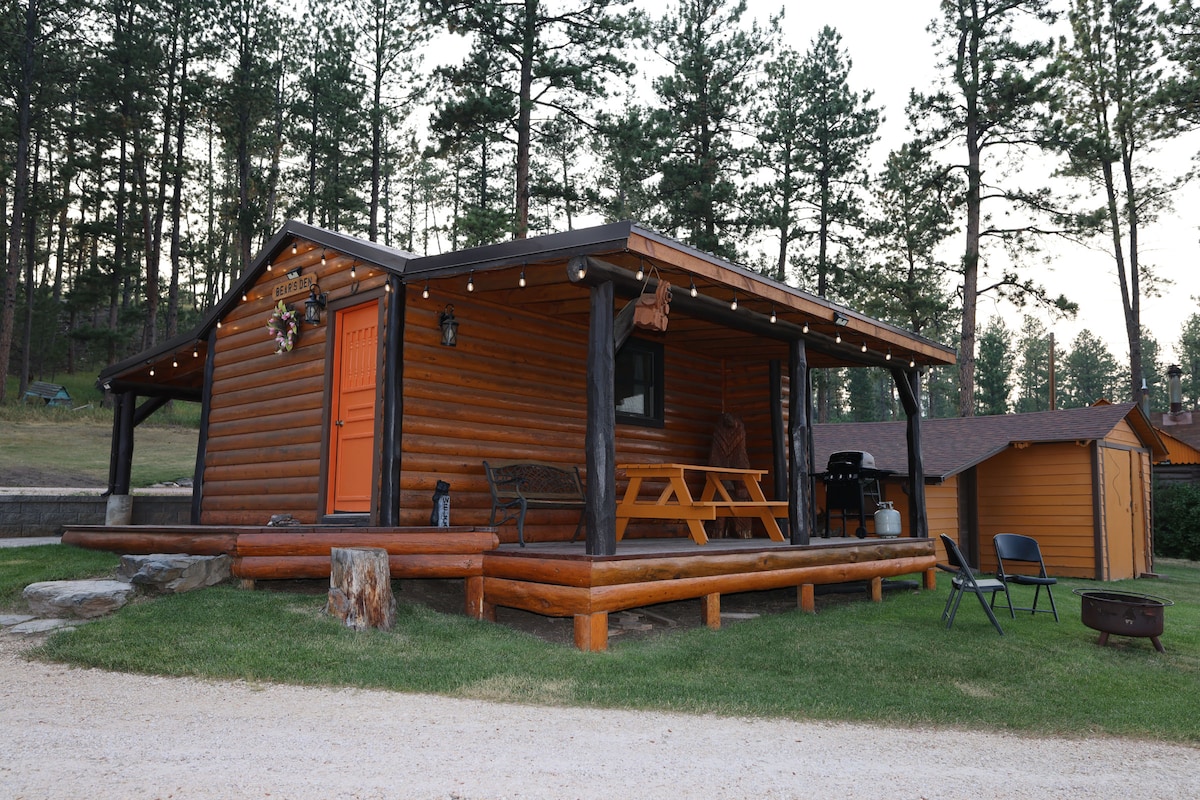 The cabin exterior features rustic wooden siding and a welcoming orange door. A picnic table is positioned on the spacious front deck, with a grill nearby. Surrounding pine trees create a serene backdrop, while a gravel pathway leads to the entrance.