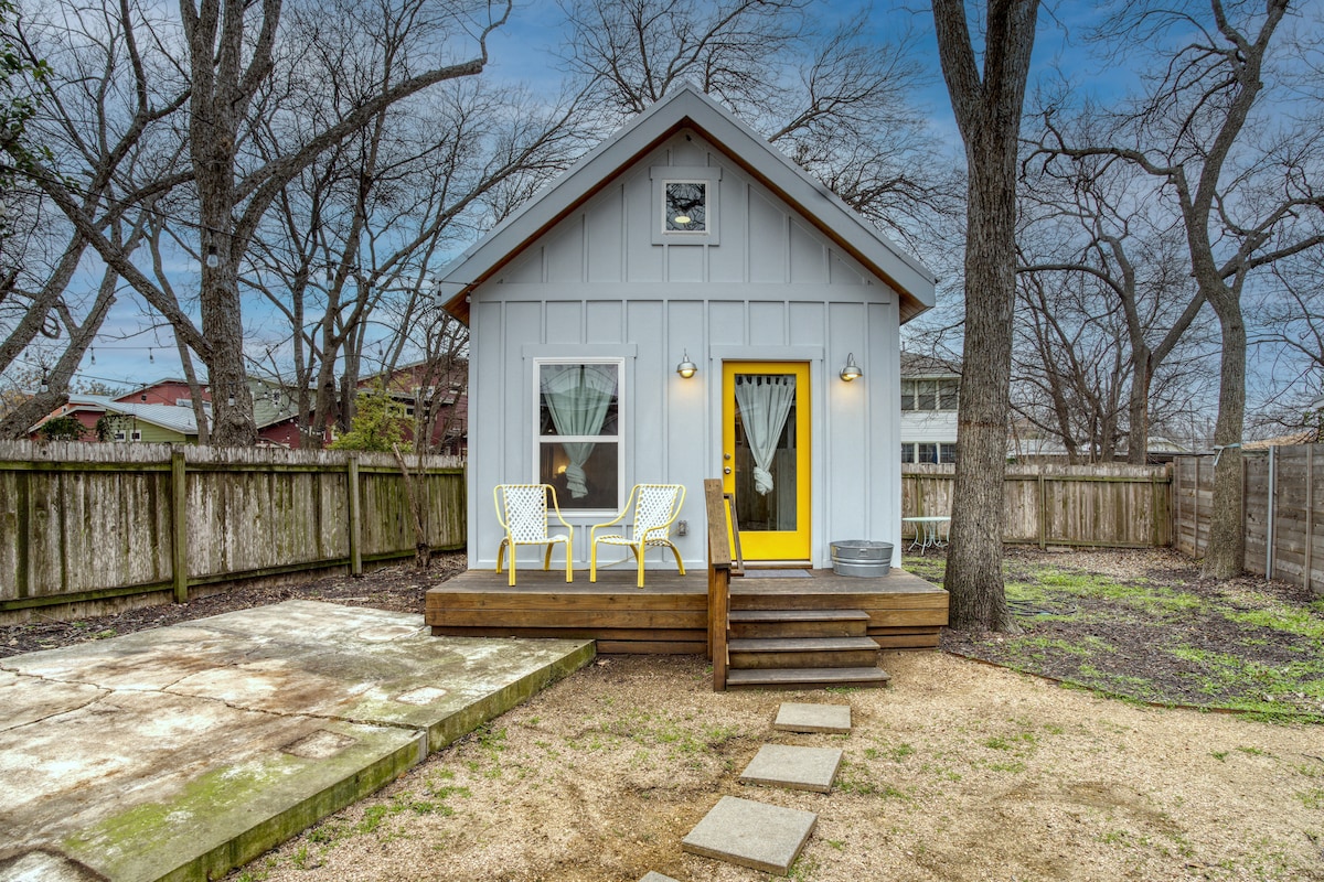 A charming backyard cottage is featured, showcasing a light blue exterior with a bright yellow door. Two white outdoor chairs are positioned on a wooden deck, surrounded by a natural landscape of trees and a gravel pathway leading to the entrance.