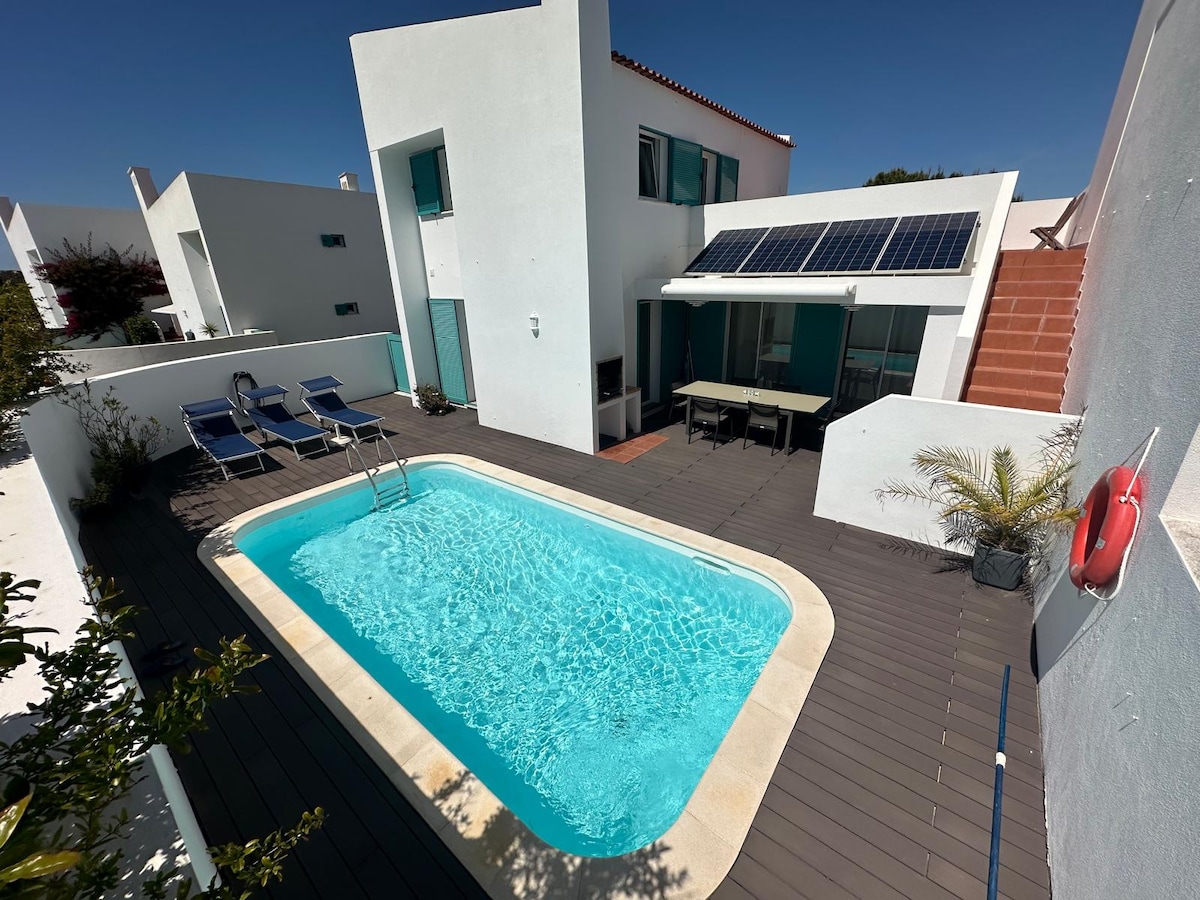 A private swimming pool is framed by a wooden deck, equipped with two lounge chairs. A spacious outdoor dining area is visible beneath a shaded section. The modern architecture of the white house features large windows and a solar panel above the patio.