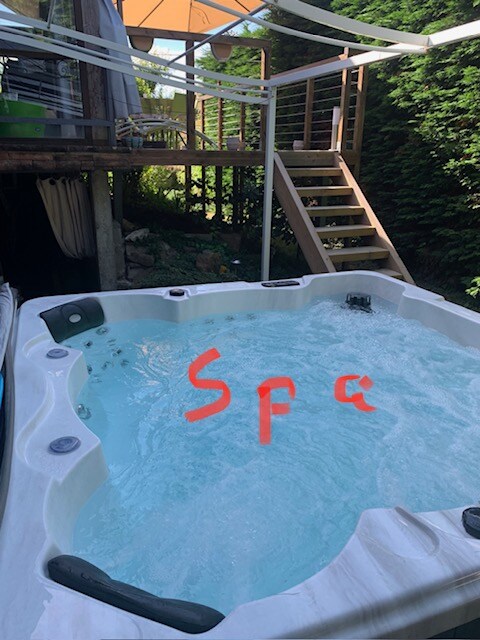 A private outdoor jacuzzi is set in a serene garden area, surrounded by lush greenery. Steps lead to a raised deck providing access to the main house. Soft water bubbles create a relaxing ambiance, inviting guests to unwind in this peaceful space.
