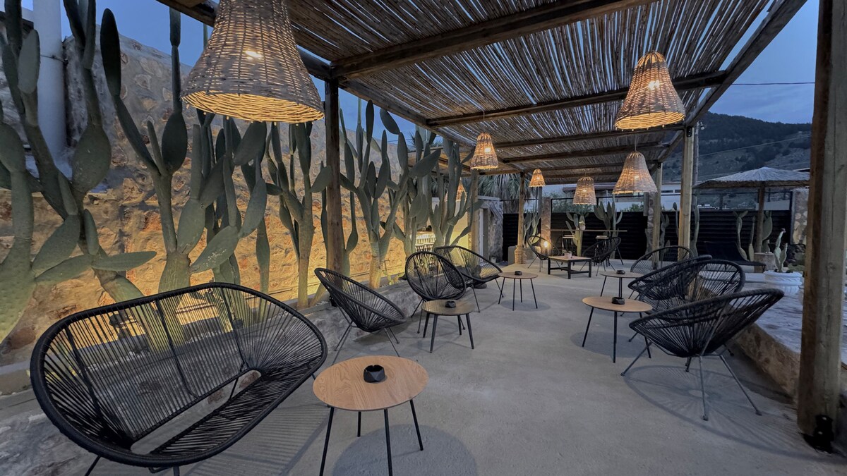 A shaded outdoor seating area is presented, featuring several black woven chairs arranged around small wooden tables. Soft lighting is provided by pendant lamps overhead, casting a warm glow on the surrounding stone walls adorned with tall, desert-like cacti.