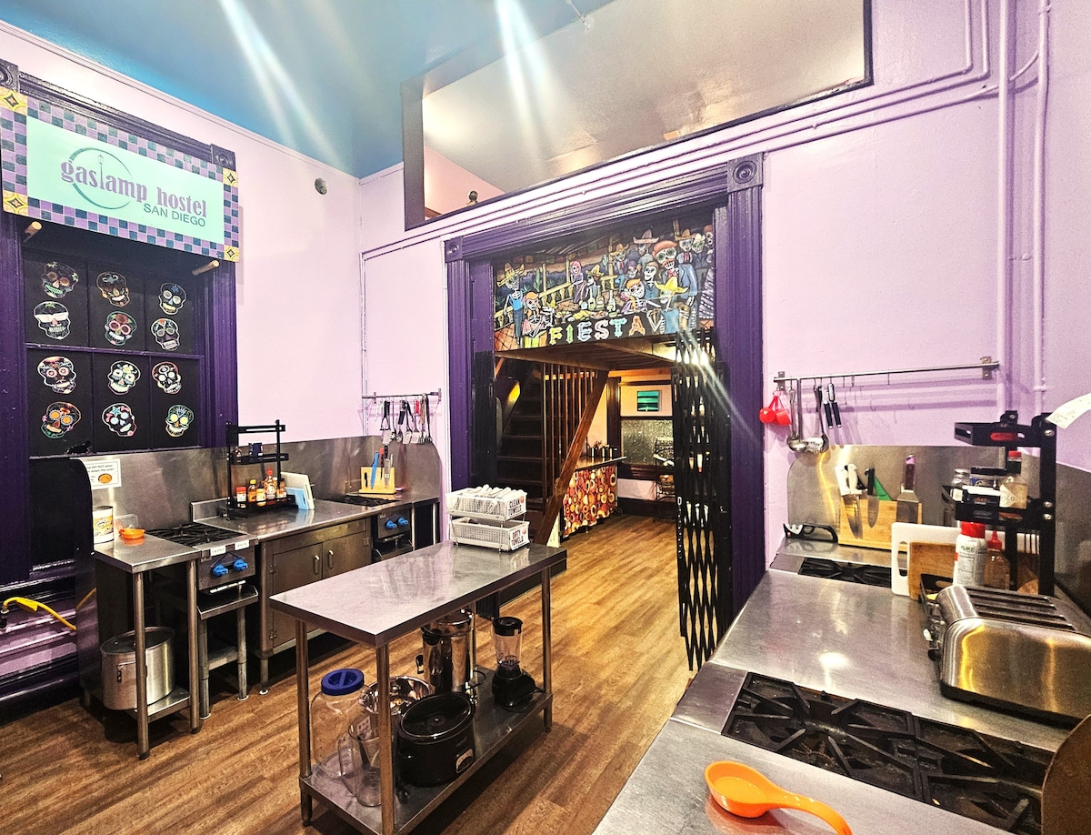 A communal kitchen area features stainless steel countertops and modern appliances. Colorful murals adorn the walls, contributing to a lively atmosphere. Shelves hold cooking utensils, and a window provides natural light to the space, enhancing accessibility for preparing meals.