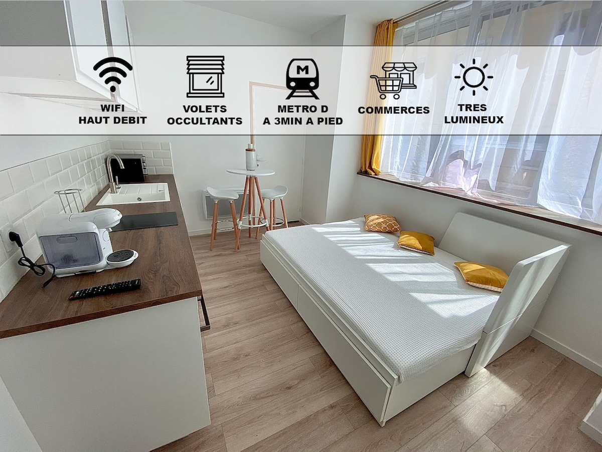A bright studio featuring a spacious bed adorned with yellow cushions, complemented by a cozy dining area with two stools. A well-equipped kitchenette is present along one wall, enhancing the functionality of the space, while natural light filters through the sheer curtains.
