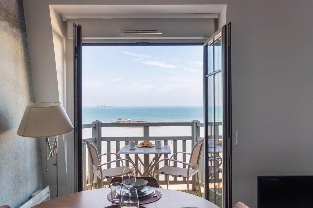 A sea-view balcony is showcased, featuring a table and two chairs. The glass doors are opened wide, allowing natural light to flood the interior and providing an unobstructed view of the tranquil ocean and distant coastline.