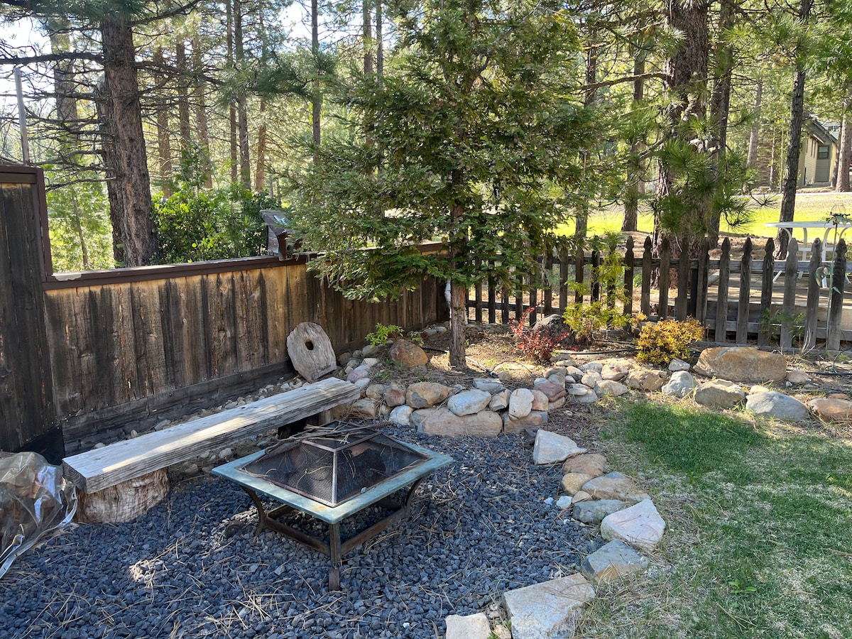 A quaint outdoor fire pit is surrounded by a circle of smooth stones, creating a cozy gathering space. Decorative landscaping features a variety of shrubs and trees, enhancing the natural setting. A rustic wooden bench is positioned nearby, inviting relaxation in the serene atmosphere.
