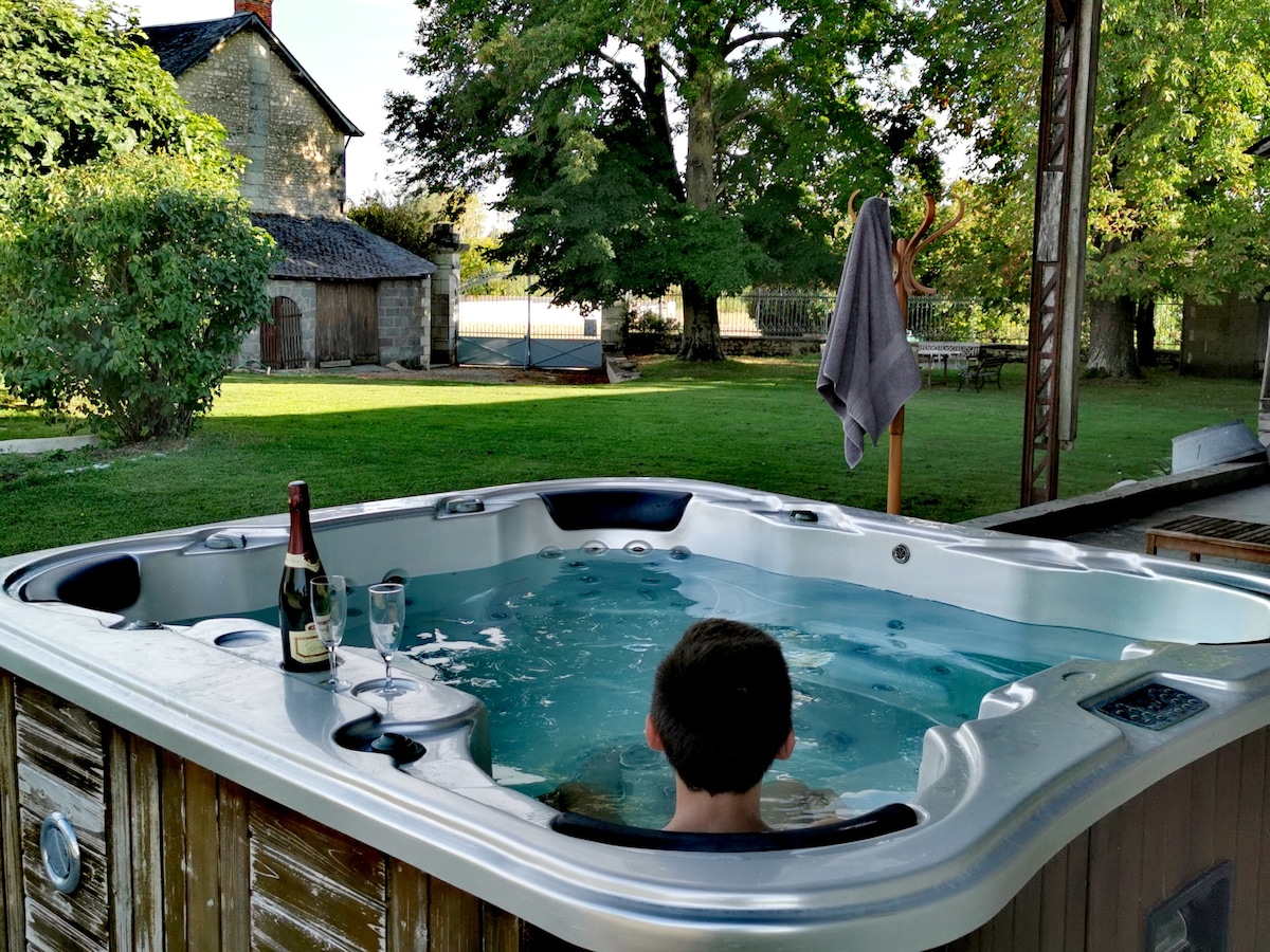 A hot tub is positioned on a covered terrace, with a person relaxing inside. The surrounding garden features lush greenery, a shaded area with a tree, and a glimpse of a rustic building in the background. A bottle of champagne and two glasses rest on the edge of the hot tub.