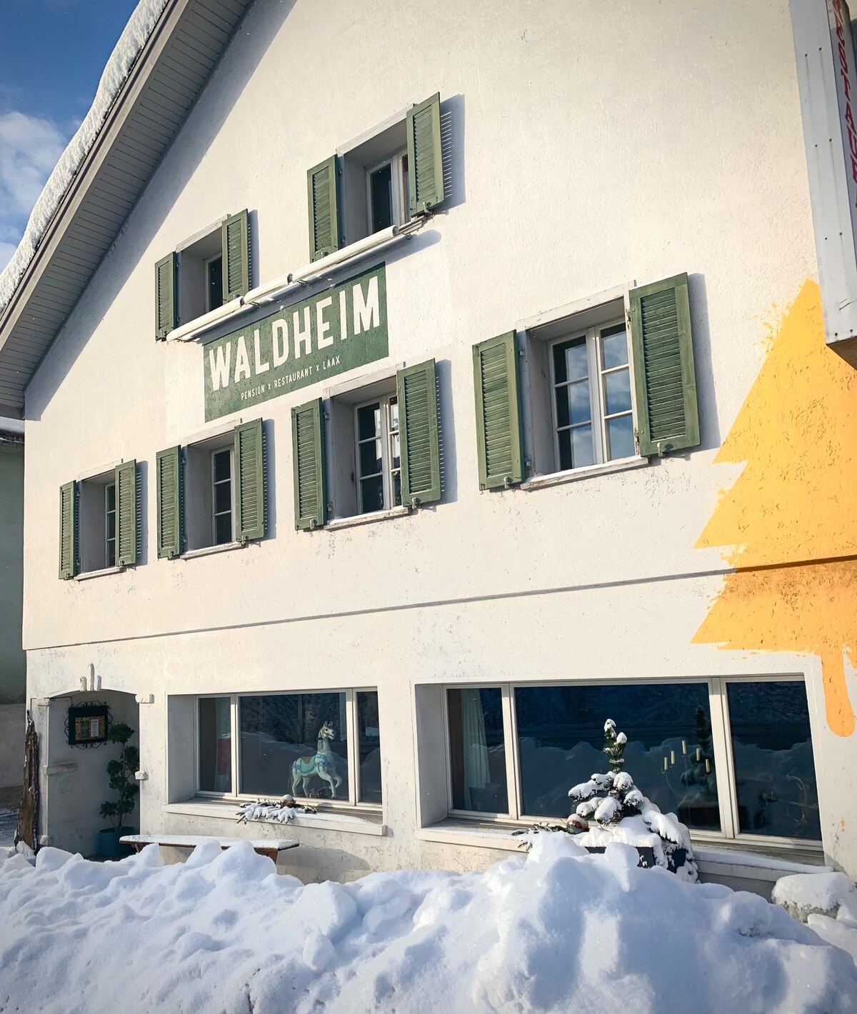 The exterior of the building features a rustic facade with green shutters and the name 'Waldheim' prominently displayed. Snow gently covers the ground and outdoor seating areas, creating a winter scene. Large windows allow glimpses of the interior while enhancing the inviting appearance.