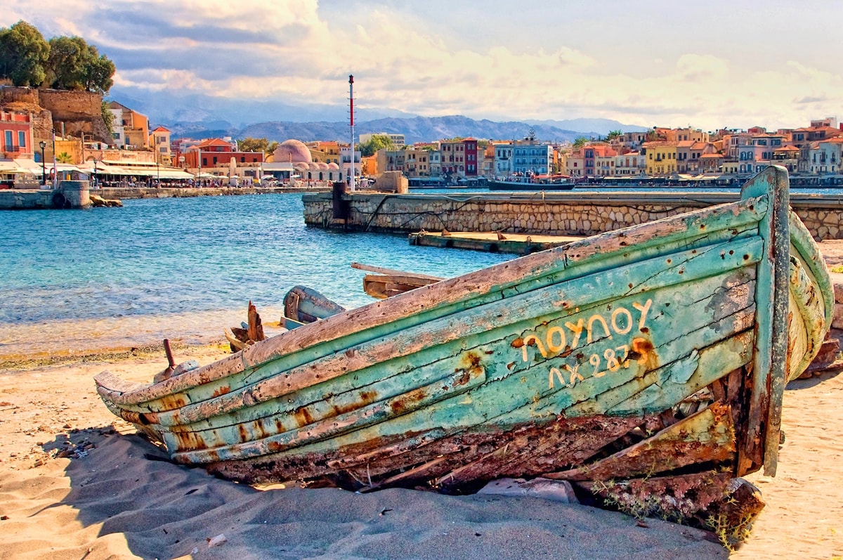 An aged wooden boat rests on sandy shore, displaying weathered green paint and faint lettering. Behind the boat, the calm blue waters stretch towards a colorful town with historic buildings, while mountains are faintly visible in the distance under a partly cloudy sky.