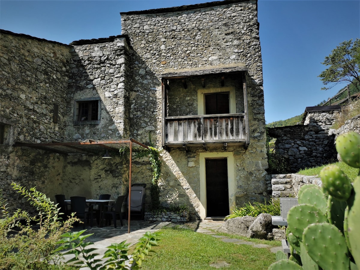 The exterior of the stone house features a rustic balcony and a shaded sitting area with a table and chairs. Green foliage surrounds the structure, and a pathway leads to the entrance. Cacti are visible in the foreground, adding a touch of greenery.
