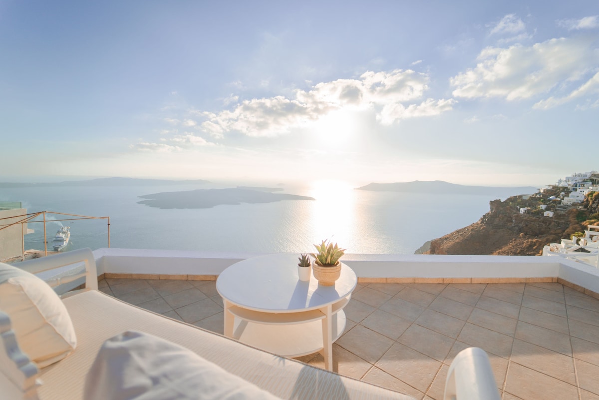 A spacious terrace offers a panoramic view of the caldera and surrounding islands, bathed in soft sunlight. A round table sits against a backdrop of the shimmering sea, while a potted plant adds a touch of greenery to the serene outdoor space.