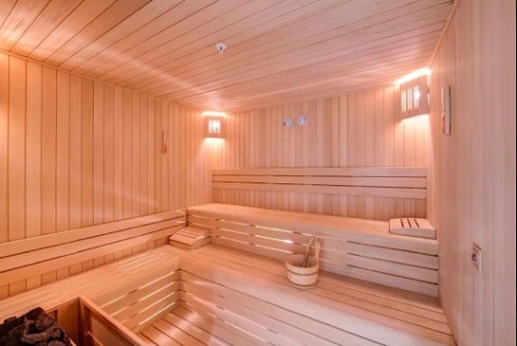 A wooden sauna interior features bench seating arranged along the walls and illuminated by soft, warm lighting. The space includes a bucket for water and a rock heater, creating a serene and relaxing environment.