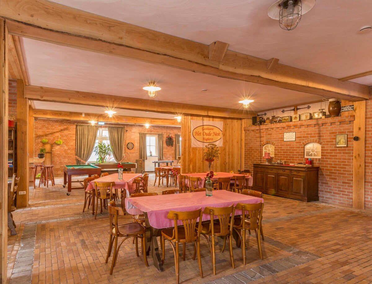 The spacious common area features wooden beams and exposed brick walls, creating a warm and inviting atmosphere. Tables with pink tablecloths are arranged for dining, complemented by wooden chairs. A pool table is visible in one corner, with natural light streaming through large windows.