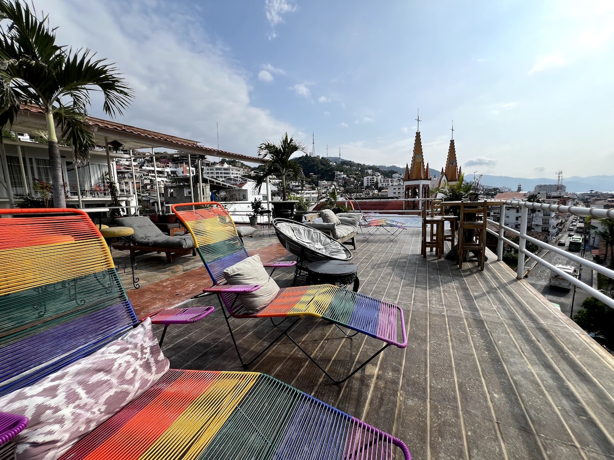 A vibrant rooftop terrace showcases colorful lounge chairs arranged for relaxation, with a backdrop of city views and distant church spires. Lush palm trees provide subtle shade, while the wooden deck enhances the open-air atmosphere, ideal for enjoying sunsets.