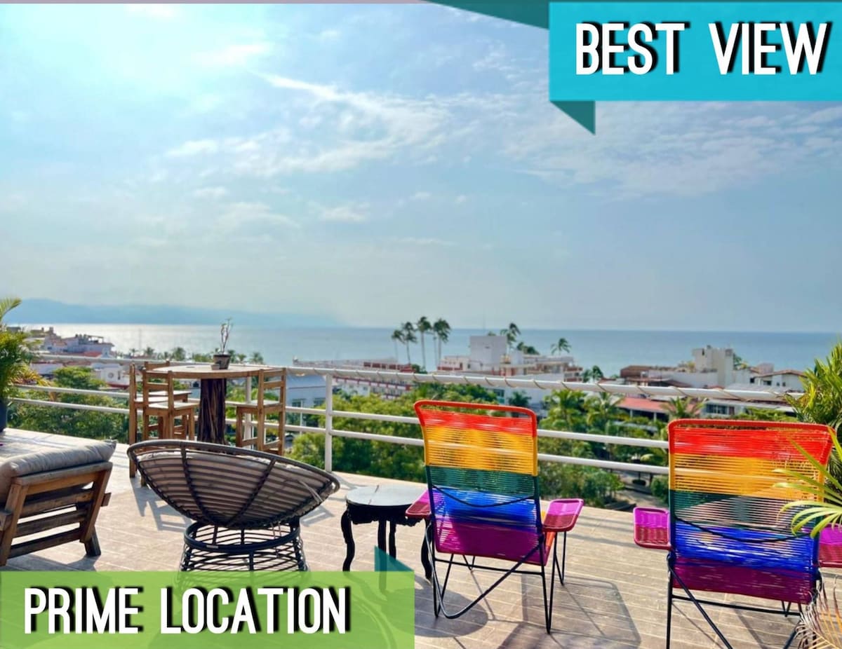 A spacious rooftop terrace features colorful chairs and a lounge area, offering expansive views of the coastline and nearby palm trees. The bright sky and distant ocean create a serene backdrop for relaxation.