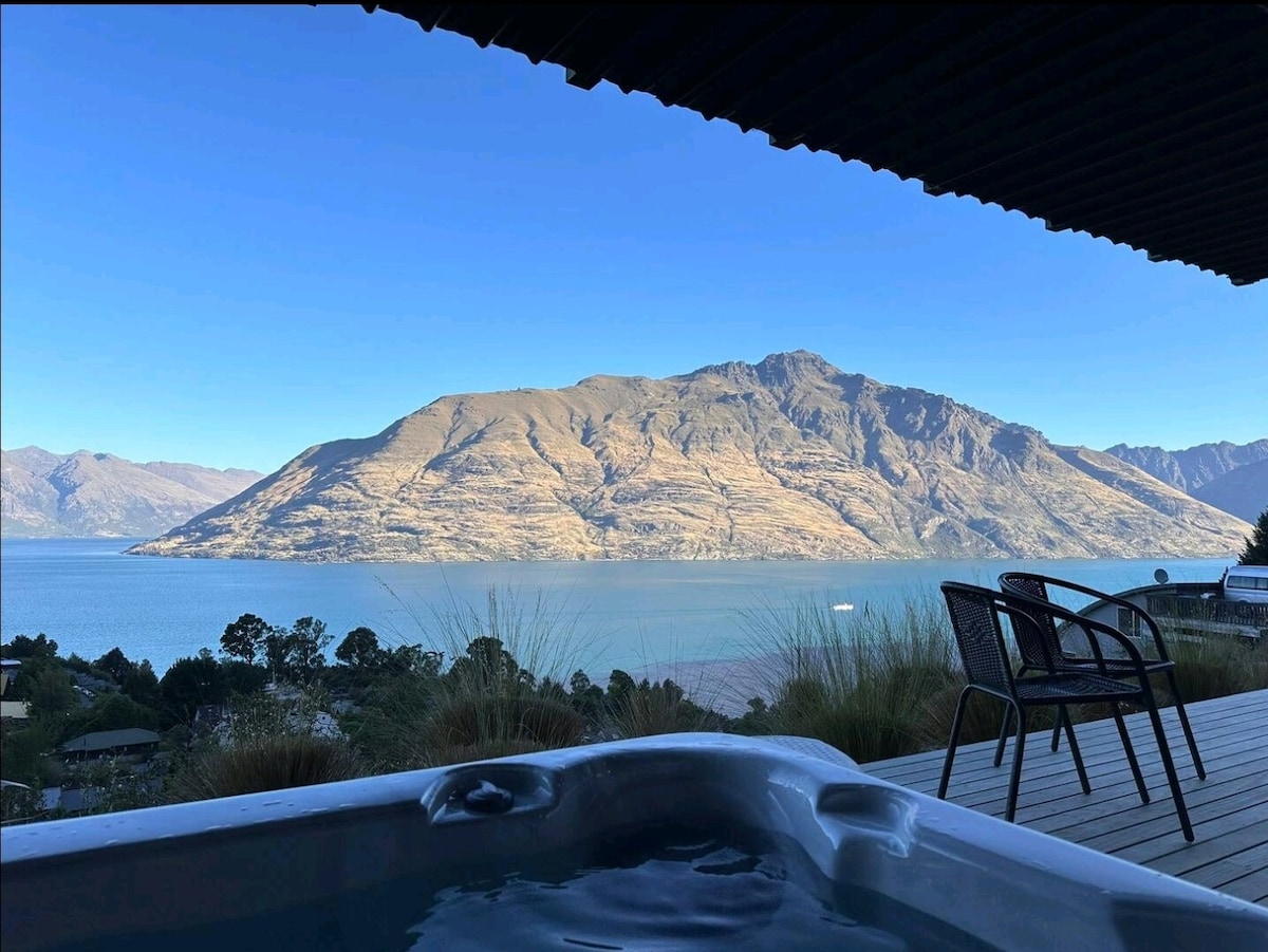 A tranquil view of Lake Wakatipu and The Remarkables is captured from the perspective of a private spa. The serene water reflects the mountains in the distance, with clear blue skies above. Two black chairs are positioned on the wooden deck, inviting relaxation.